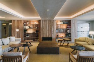 El moderno salón del Hotel Chiqui, ideal para una Escapada Romántica en el hotel Chiqui (Santander), cuenta con estanterías de madera, lujosos sofás, mesas redondas y una suave iluminación cenital en tonos cálidos con un diseño minimalista para un ambiente tranquilo. - ForTwo - Planes para parejas
