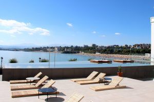 La piscina infinita en la azotea del Hotel Chiqui ofrece impresionantes vistas a la playa y al paisaje urbano de Santander, ideal para una "Escapada romántica en el hotel Chiqui (Santander)", con tumbonas bajo un cielo azul claro. - ForTwo - Planes para parejas