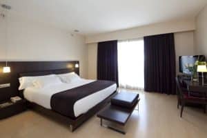 Disfrute de la tranquilidad en una moderna habitación en Escapada con spa en el hotel HM Jaime III, con una cama grande, muebles de madera oscura y televisor. Relájese junto a la ventana del balcón con cortinas y descanse a solo unos minutos del lujoso spa en la vibrante Palma de Mallorca. - ForTwo - Planes para parejas