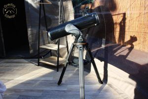 En una habitación soleada con suelo de madera, un telescopio sobre un trípode sugiere aventuras astronómicas similares a la "Escapada romántica en hotel burbuja con Gredos Estelar" en Ávila. Cerca, una escalera de dos peldaños se yergue, como invitando al cielo nocturno de Ávila a entrar. - ForTwo - Planes para parejas