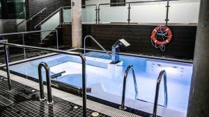 Experimente la Escapada 50 Sombras de Gris con Spa, que cuenta con un jacuzzi privado con pasamanos metálicos, escalones y salvavidas. Las mamparas de cristal ofrecen privacidad mientras el agua azulada crea un ambiente de spa. - ForTwo - Planes para parejas