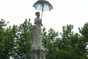 En un rincón sereno de Barcelona, el "Free tour de las mujeres ilustres de Barcelona" presenta una elegante estatua de una mujer con un paraguas, rodeada de frondosos árboles y contra un cielo nublado. - ForTwo - Planes para parejas