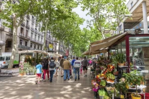 Paseando por un mercado callejero arbolado en Barcelona, te encontrarás con coloridos puestos de flores junto a edificios de varias plantas, que reflejan el vibrante espíritu de la ciudad. Recuerda al "Tour gratuito de las mujeres ilustres de Barcelona", que celebraba a las mujeres que antaño paseaban por estas avenidas. - ForTwo - Planes para parejas