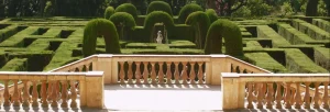 Una elegante terraza de piedra con vistas a un exuberante laberinto de setos que recuerda al Laberinto de Horta de Barcelona, con una cautivadora escultura central. Disfrute de la belleza con nuestro "Tour gratuito por el Laberinto de Horta (Barcelona)". - ForTwo - Planes para parejas