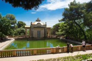 Descubra una experiencia pintoresca en el Laberinto de Horta, Barcelona, donde un antiguo pabellón junto a una tranquila piscina se baña de luz natural, rodeado de frondosos árboles y una valla decorativa. Disfrute de una visita inolvidable con el "Tour gratuito por el Laberinto de Horta (Barcelona)". - ForTwo - Planes para parejas