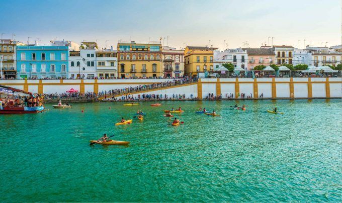 Los kayakistas se deslizan por un ancho río, mientras el vibrante barrio de Triana de Sevilla, protagonista de la experiencia 'Tour gratuito por el barrio de Triana', y los espectadores se alinean en la orilla al fondo. - ForTwo - Planes para parejas