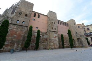 Un edificio histórico de piedra con arco, enclavado entre altos árboles y con el cielo nublado como fondo, captura a la perfección la esencia de la antigua Barcelona, ideal para el "Tour gratuito por la Barcelona romana". - ForTwo - Planes para parejas