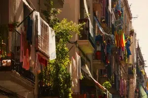 La ropa en tonos vivos se balancea en los balcones iluminados por el sol a lo largo de una estrecha calle de Barcelona, con plantas vibrantes decorando las barandillas, una vista común que puede encontrar en un tour gratuito por Barcelona. - ForTwo - Planes para parejas