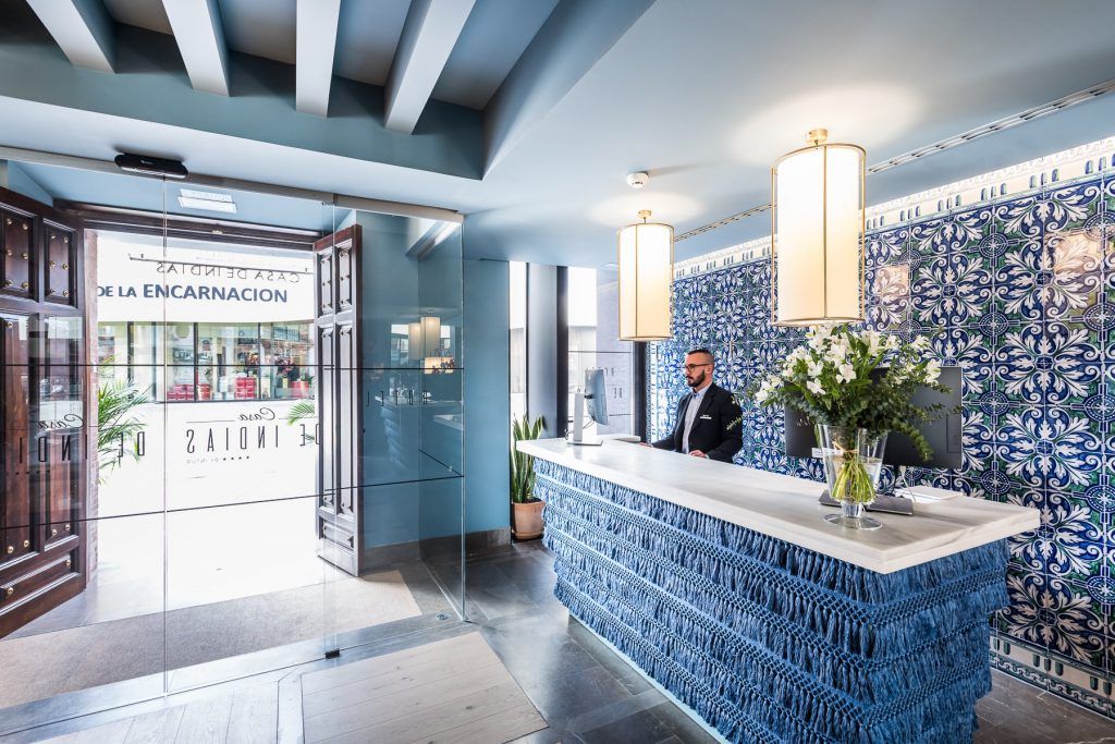 En el Hotel Casa de Indias By Intur de Sevilla, un hombre se encuentra en el mostrador de recepción blanco sobre un fondo de azulejos azules con flores vibrantes, cerca de la tranquila entrada del spa. Disfrute de una estancia con paquete de spa y desayuno en este tranquilo entorno. - ForTwo - Planes para parejas