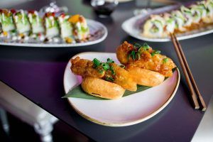 Los bocadillos de pollo frito sobre panecillos con guarnición verde ocupan un lugar central, mientras que los deliciosos rollitos de Miss Sushi Granada permanecen en el fondo, ofreciendo una muestra de la vibrante esencia de Granada. - ForTwo - Planes para parejas