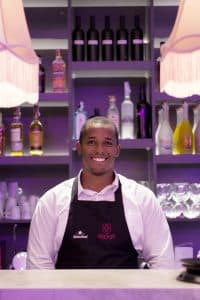Un camarero con camisa blanca y delantal negro sonríe detrás de la barra en Miss Sushi Granada, con una vibrante exhibición de botellas de licor y vasos. - ForTwo - Planes para parejas