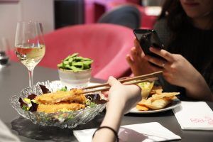 En Miss Sushi Granada, uno usa palillos para disfrutar la comida de un plato mientras otro captura el momento con su teléfono; el vino y los deliciosos bocadillos en la mesa realzan la inolvidable experiencia culinaria. - ForTwo - Planes para parejas