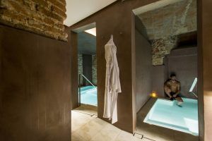 Una persona se relaja en una zona de spa con piscina con paredes de ladrillo visto y un albornoz colgado cerca, perfecta para una Escapada con desayuno y spa en Balneario Caldes de Montbui (Barcelona). - ForTwo - Planes para parejas