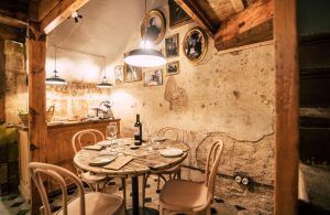 Un acogedor comedor rústico cuenta con una mesa redonda y cuatro sillas, complementadas con una botella de vino, que evoca el cálido ambiente de una comida barcelonesa. Lámparas colgantes y fotos vintage realzan el encanto inspirado en la "Comida o cena de productos locales en l'Espai Quera (Barcelona)". - ForTwo - Planes para parejas