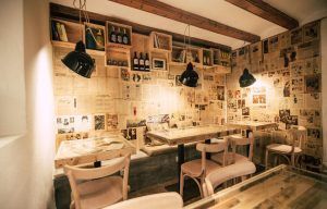 Espai Quera, una acogedora cafetería en Barcelona, cuenta con muebles de madera, paredes revestidas de periódicos y luces colgantes para un ambiente vintage mientras ofrece "Comida o cena de productos locales" para disfrutar. - ForTwo - Planes para parejas