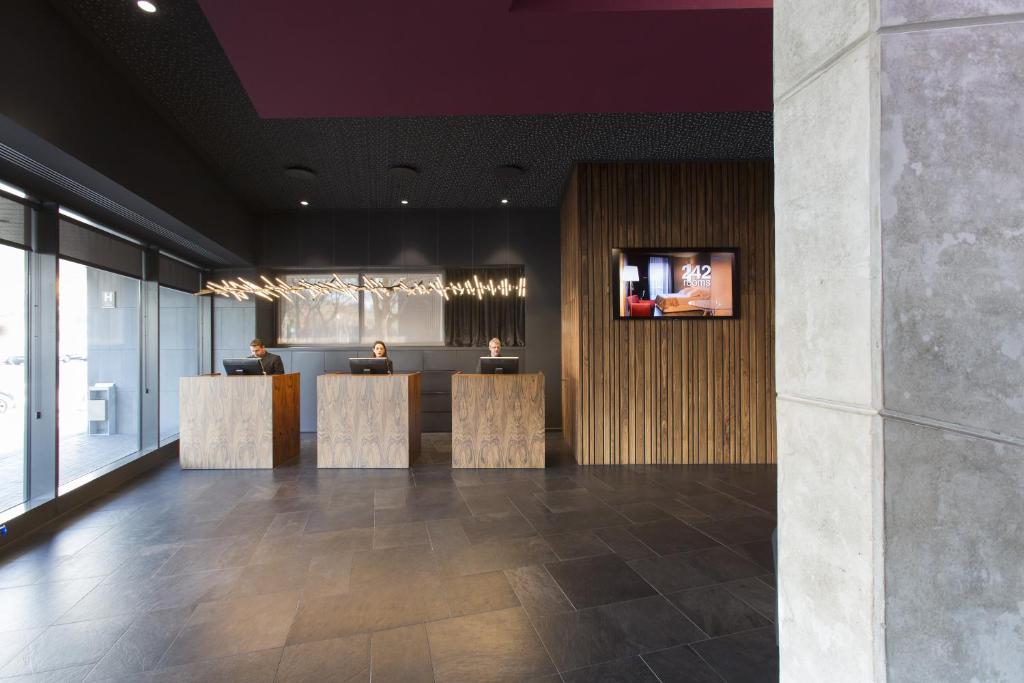 El hotel con jacuzzi SB Plaza Europa de Barcelona cuenta con un moderno vestíbulo con tres mostradores de facturación, detalles en madera y una pantalla montada en la pared, preparando el escenario para una estancia de lujo que incluye relajarse en un jacuzzi. - ForTwo - Planes para parejas