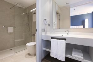 Disfrute de un baño moderno en el Hotel con jacuzzi SB Plaza Europa en Barcelona, con una elegante ducha de cristal, lavabo blanco, secador de pelo montado en la pared, cómodas toallas en el estante y estante, y disfrute de un exquisito jacuzzi para una relajación máxima. - ForTwo - Planes para parejas