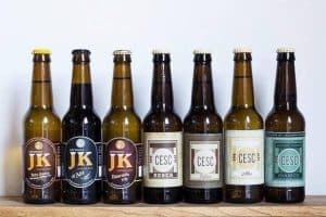 Siete botellas de cerveza surtidas de La Cervecería de Poblenou, cada una con etiquetas diferentes, se encuentran sobre una superficie de madera, capturando el espíritu vibrante del Poblenou de Barcelona sobre un fondo liso. - ForTwo - Planes para parejas