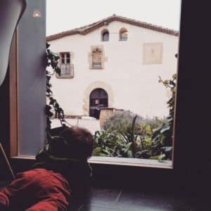 Un niño vestido de rojo mira por una ventana hacia Can Calopa, conocido por "Enoturismo con cata en Can Calopa con vistas desde Collserola (Barcelona)", elegantemente ubicado entre una exuberante vegetación. - ForTwo - Planes para parejas