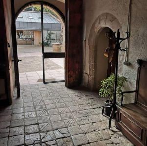 El pasillo con suelo de piedra, con puerta arqueada, perchero y planta, conduce al encantador patio del Hotel Lotus Priorat en Falset. Un cartel de LA VINOTECA sugiere aventuras enoturísticas más allá, con la frase "Escapada de enoturismo de la mano del hotel Lotus Priorat". - ForTwo - Planes para parejas