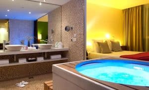 Disfrute de una estancia de lujo en el Hotel con jacuzzi SB Diagonal Zero en Barcelona, que cuenta con una habitación con bañera de hidromasaje, lavabos dobles, espejo grande, cama con colchón amarillo e iluminación ambiental, perfecta para relajarse. - ForTwo - Planes para parejas
