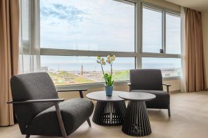 En una sala con amplios ventanales, dos sillas grises y mesas pequeñas ofrecen vistas al océano desde el SB Diagonal Zero. Una orquídea en maceta sobre una mesa añade elegancia, evocando el ambiente de un hotel chic de Barcelona. - ForTwo - Planes para parejas