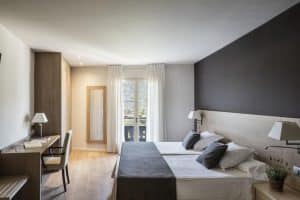 Una moderna habitación de hotel con dos camas individuales, escritorio, silla, un gran ventanal con cortinas blancas y decoración en tonos marrones y blancos ofrece una escapada tranquila. Disfrute del spa cercano con aguas termales rejuvenecedoras reservando el "Pack noche + spa en el paraíso más exclusivo de agua termal milenaria" en Barcelona. - ForTwo - Planes para parejas