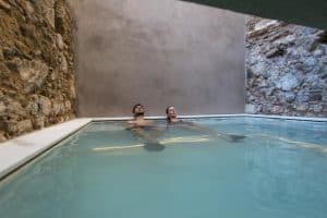 Dos personas se relajan en una estrecha piscina cubierta con paredes de piedra y hormigón, disfrutando del ambiente de una noche de spa con el "Pack noche + spa en el paraíso más exclusivo de agua termal milenaria (Barcelona). - ForTwo - Planes para parejas