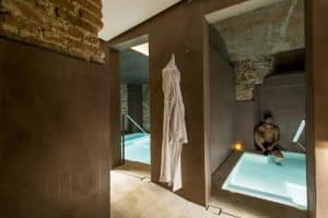 En el sereno resplandor del Pack Noche + Spa en el Paraíso Más Exclusivo de Agua Termal Milenaria en Barcelona, un hombre se relaja en una moderna piscina cubierta de spa con una lujosa bata blanca colgando cerca. - ForTwo - Planes para parejas