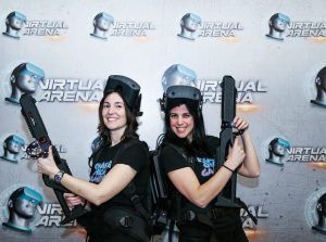 Dos mujeres sonríen con cascos y controladores de realidad virtual, de pie frente a un telón de fondo de Virtual Arena, listas para la Experiencia de Realidad Virtual Multijugador en Virtual Arena (Madrid). - ForTwo - Planes para parejas