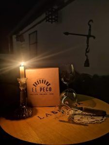 Se desarrolla una escena a la luz de las velas con una caja de vino, una copa y un cigarro Degustación de vinos naturales en Destino Starlight Cielos de Sierra Morena elegantemente dispuestos sobre la mesa, proyectando suaves sombras en la pared. - ForTwo - Planes para parejas