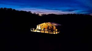 Una carpa iluminada en medio de un paisaje oscuro bajo un cielo crepuscular con árboles recortados captura el encanto sereno de la Cabaña junior suite con piscina privada en La Dehesa Experiences (Córdoba). - ForTwo - Planes para parejas