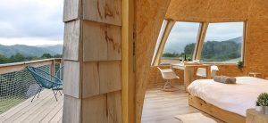Un acogedor dormitorio rústico en Navarra con grandes ventanales ofrece vistas a la montaña que recuerdan a los Iglús panorámicos en plena Selva de Irati y cuenta con una terraza de madera con una silla. - ForTwo - Planes para parejas
