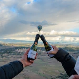 Dos personas brindan con botellas al amanecer mientras un globo aerostático sobrevuela el pintoresco paisaje de Mallorca. Experimente el "Vuelo en globo privado para dos al amanecer (Mallorca)". - ForTwo - Planes para parejas
