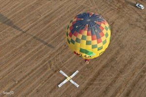Un colorido globo aerostático sobrevuela un campo con un objetivo a sus pies, capturando la serena belleza de Mallorca. Cerca, un vehículo privado espera a los aventureros tras su fascinante experiencia de "Vuelo en globo privado para dos al amanecer (Mallorca)". - ForTwo - Planes para parejas