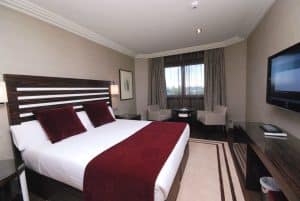 Disfrute de máxima privacidad en Madrid con el Hotel Los Peñascales. Esta elegante habitación cuenta con una cama grande, detalles en burdeos, un televisor de pantalla plana y dos sillas junto a la ventana, combinando a la perfección comodidad y decoración moderna. - ForTwo - Planes para parejas
