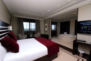Disfrute de la máxima privacidad en el Hotel Los Peñascales de Madrid. Esta habitación ofrece una cama doble con almohadas y cubrecamas color granate, un amplio ventanal y un diseño diáfano con bañera, creando un oasis de confort. - ForTwo - Planes para parejas