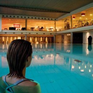 Una persona en una piscina contempla el interior de un edificio elegante e iluminado con grandes ventanales y zonas para sentarse, que recuerda a Almuerzo, Circuito de Spa y habitación romántica con Cava y Fresas (Barcelona). - ForTwo - Planes para parejas