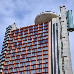 Un moderno edificio de gran altura con sus singulares ventanas de rejilla roja y su estructura superior circular hace eco de las curvas icónicas de Almuerzo, Circuito de Spa y habitación romántica con Cava y Fresas (Barcelona), frente a un cielo azul. - ForTwo - Planes para parejas