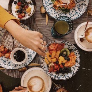 Vista superior de una mesa en el Gran Hotel Inglés con fruta, cruasanes y bebidas. Una mano toma un trozo con un tenedor mientras saborea el cóctel con acceso a jacuzzi y masaje en el Gran Hotel Inglés (Madrid) cercano. - ForTwo - Planes para parejas