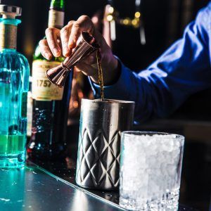 En el Gran Hotel Inglés de Madrid, el barman prepara con maestría un cóctel en la elegante barra, rodeado de relucientes botellas y un vaso de hielo. Disfrute de su bebida mientras disfruta del acceso al jacuzzi y a los servicios de masajes. - ForTwo - Planes para parejas