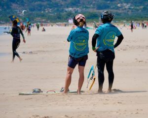 Dos personas con camisetas azules y cascos se encuentran en la playa de Tarifa con el Curso de KiteSurf para dos personas de Subcielo KiteSchool, mirando a otros practicar kitesurf. - ForTwo - Planes para parejas
