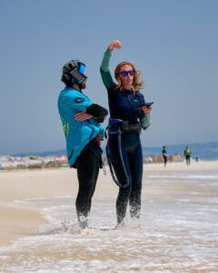 Dos personas en una playa con trajes de neopreno y cascos, una de ellas haciendo un gesto hacia arriba con la impresionante costa de Tarifa al fondo, parecen listas para una aventura con el Curso de KiteSurf para dos personas de Subcielo KiteSchool Tarifa (Cádiz). - ForTwo - Planes para parejas