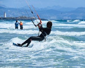 En un día soleado en Tarifa, un kitesurfista navega por las olas cerca de un pintoresco faro, acompañado por otros entusiastas del "Curso de KiteSurf para dos personas" de Subcielo KiteSchool, creando un vibrante paisaje marino. - ForTwo - Planes para parejas