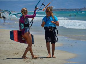 En una playa de arena de Tarifa, dos personas con material de kitesurf charlan animadamente sobre su día en el "Curso de KiteSurf para dos personas" de Subcielo KiteSchool. - ForTwo - Planes para parejas