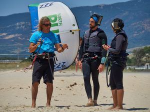 Dos entusiastas aprendices están tomando el "Curso de KiteSurf para dos personas por Subcielo KiteSchool Tarifa (Cádiz)" en una playa de arena en Tarifa, con una cometa vibrante volando de fondo. - ForTwo - Planes para parejas