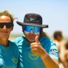 Dos personas con camisas azules y gafas de sol están afuera en un día soleado. Una levanta el pulgar, quizás celebrando una gran sesión con el emocionante "Curso de KiteSurf para dos personas" de Subcielo KiteSchool en Tarifa (Cádiz). - ForTwo - Planes para parejas