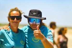Dos personas con camisas azules y gafas de sol están afuera en un día soleado. Una levanta el pulgar, quizás celebrando una gran sesión con el emocionante "Curso de KiteSurf para dos personas" de Subcielo KiteSchool en Tarifa (Cádiz). - ForTwo - Planes para parejas