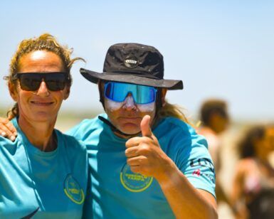Dos personas con camisas azules y gafas de sol están afuera en un día soleado. Una levanta el pulgar, quizás celebrando una gran sesión con el emocionante "Curso de KiteSurf para dos personas" de Subcielo KiteSchool en Tarifa (Cádiz). - ForTwo - Planes para parejas
