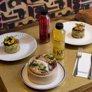 Tres platos con bebidas y botellas de kombucha sobre una mesa, complementados con una servilleta de la marca Flax & Kale, crean un entorno ideal para 'Almuerzo, Spa y Masaje con aceites veganos en hotel Yurbban (Barcelona)'. - ForTwo - Planes para parejas
